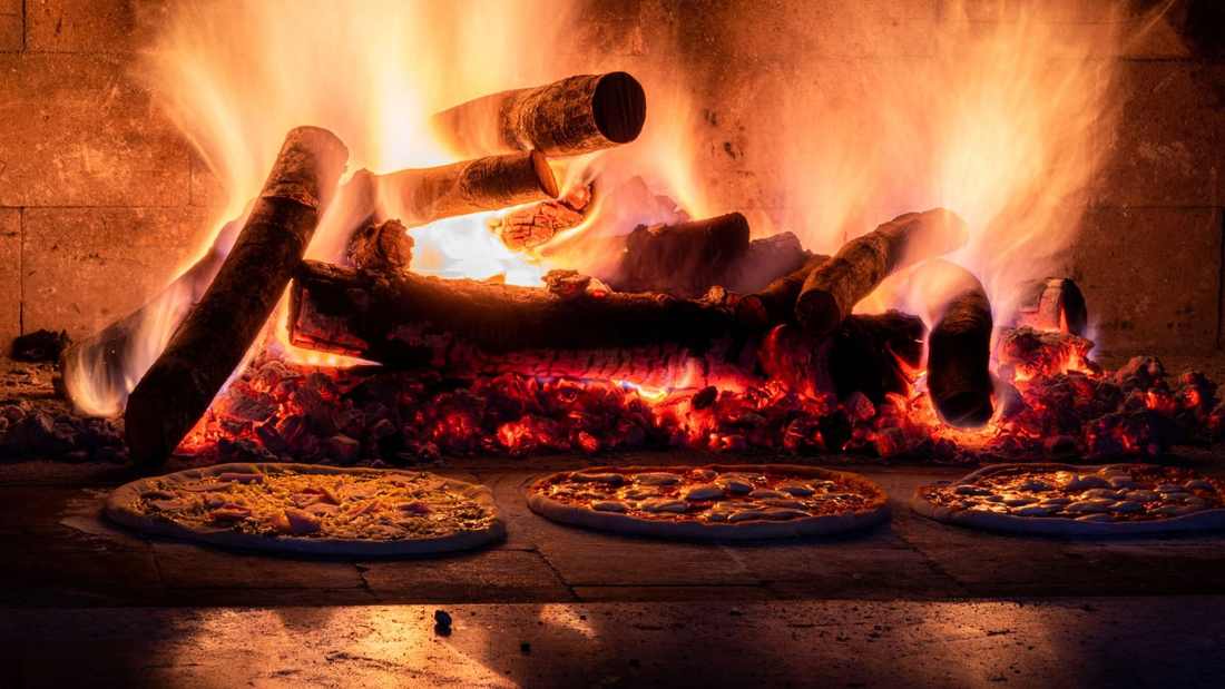Four à bois en pleine chauffe, bûches enflammées au-dessus de pizzas en cuisson